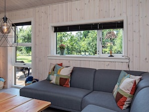A modern living room with a grey sofa, colorful cushions, a wooden table, and a window with a view of greenery. - 5 Star Holiday Home in Vaeggerlose (Vaeggerlose)