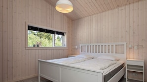 A bedroom with a wooden paneled wall, a bed with a white headboard, a bedside table, and a window with blinds. - 5 Star Holiday Home in Vaeggerlose (Vaeggerlose)