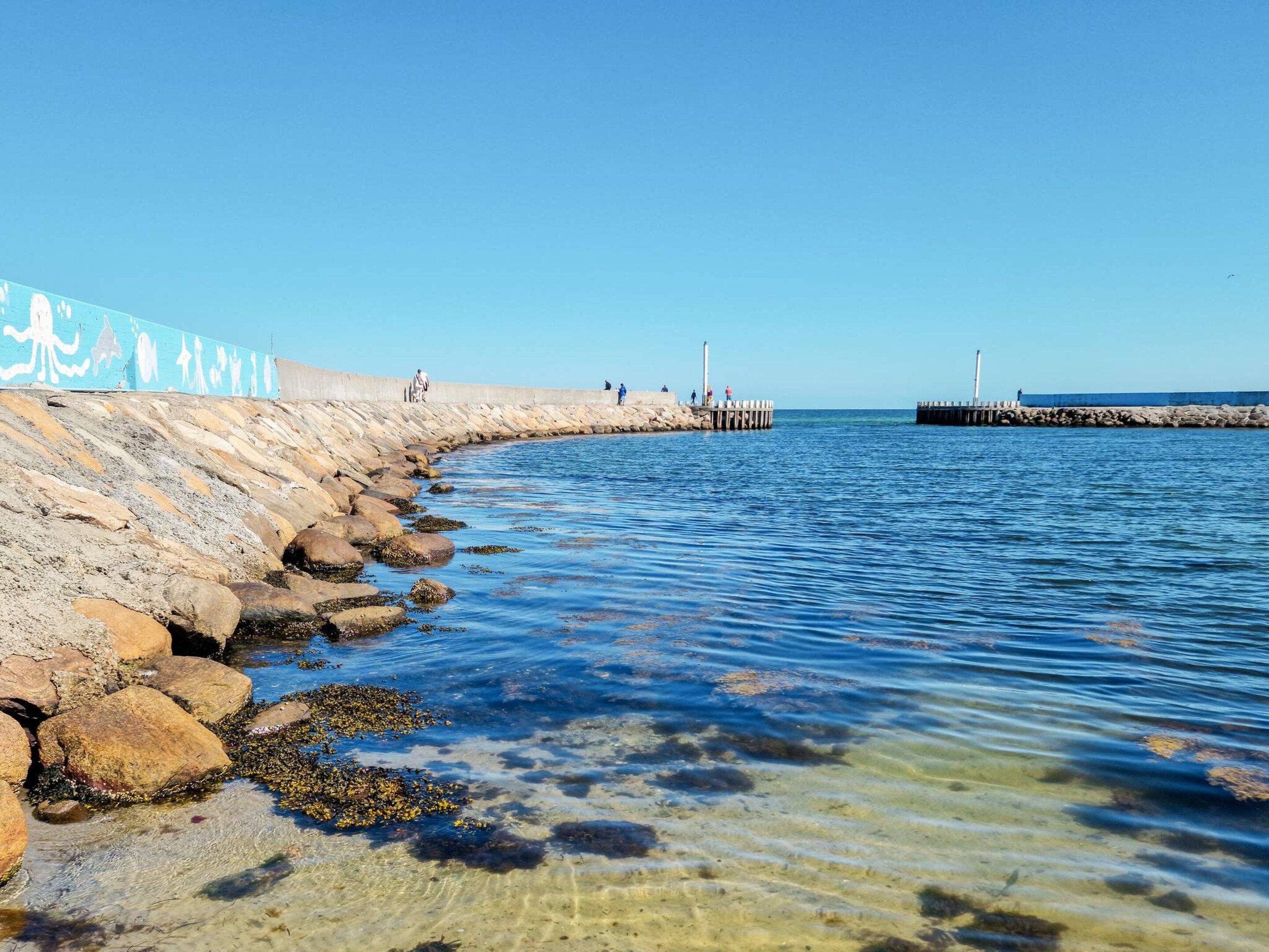 A coastal walkway with a clear view of the sea, a rocky shoreline, and a pier extending into the water.
