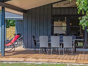 A covered outdoor seating area with a table and chairs, a wooden deck, and a view of the interior through large windows.