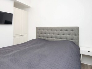 A bed with a grey upholstered headboard in a room with white walls and a built-in TV.
