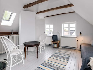 A loft-style room with a sloped ceiling, white walls, a wooden floor, a small round table, two white chairs, a television on a wooden stand, a floor lamp, and a window with a view of the outside.