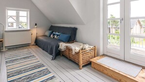 Camera mansarda con soffitto spiovente, un letto con cuscini blu, un cesto di vimini e vista attraverso un'ampia porta.