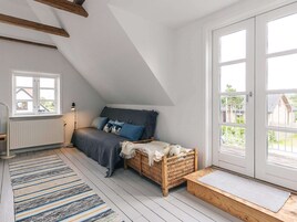 Dormitorio tipo loft con techo inclinado, una cama con almohadas azules, una cesta de mimbre y vistas a través de un gran ventanal.