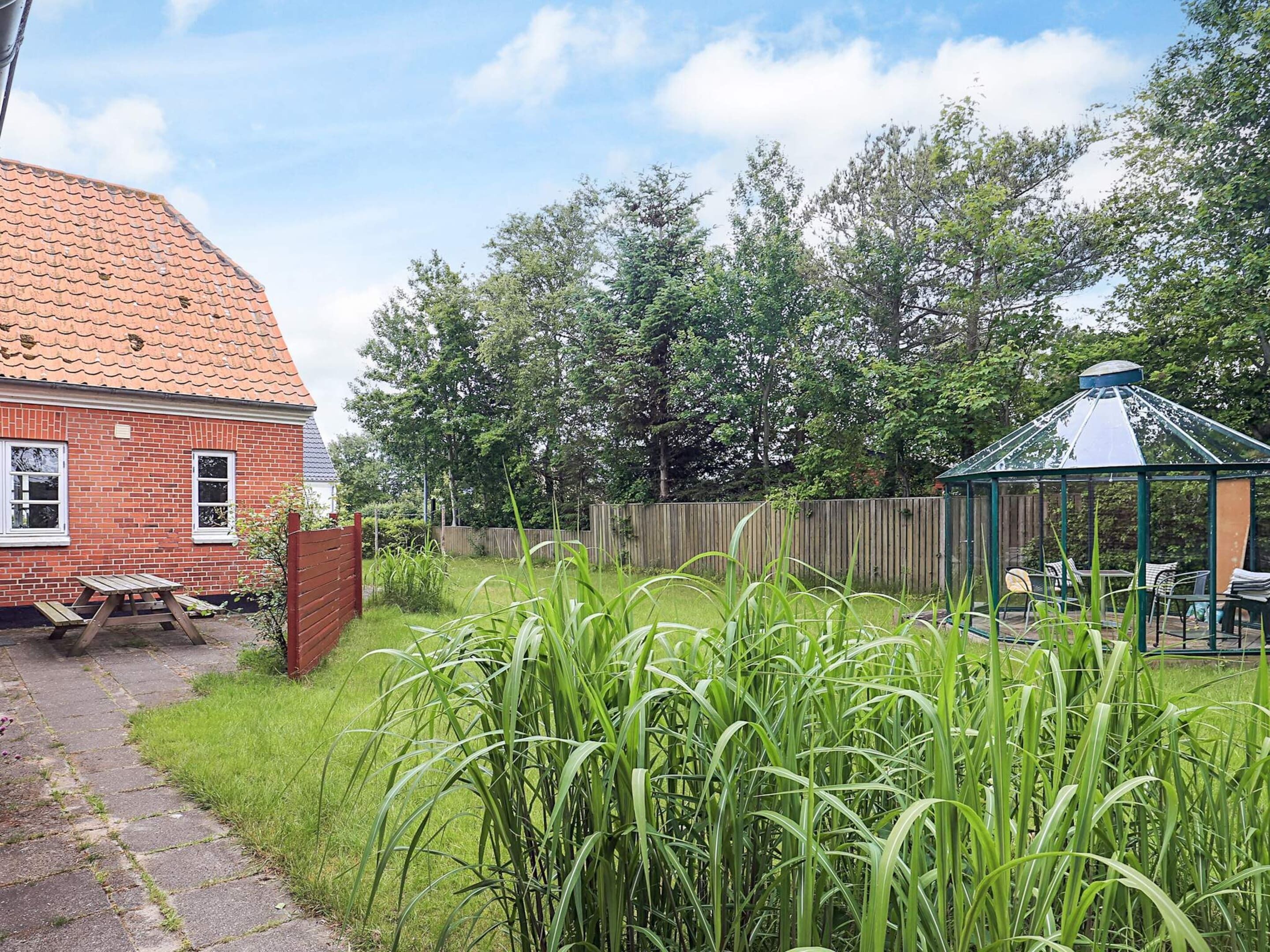 A brick house with a tiled roof, a wooden fence, and a garden with tall grass.