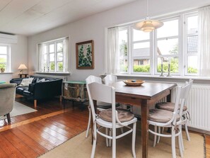 A dining area with a wooden table and white chairs, a black leather sofa, a rustic chest of drawers, and a painting on the wall.
