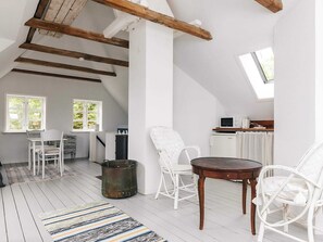 A loft-style room with exposed wooden beams, a dining table, white chairs, a small kitchen area, and a window providing natural light.