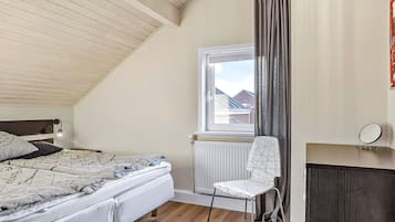 A bedroom with a sloped ceiling, a bed with patterned bedding, a chair, a window with curtains, and a radiator.