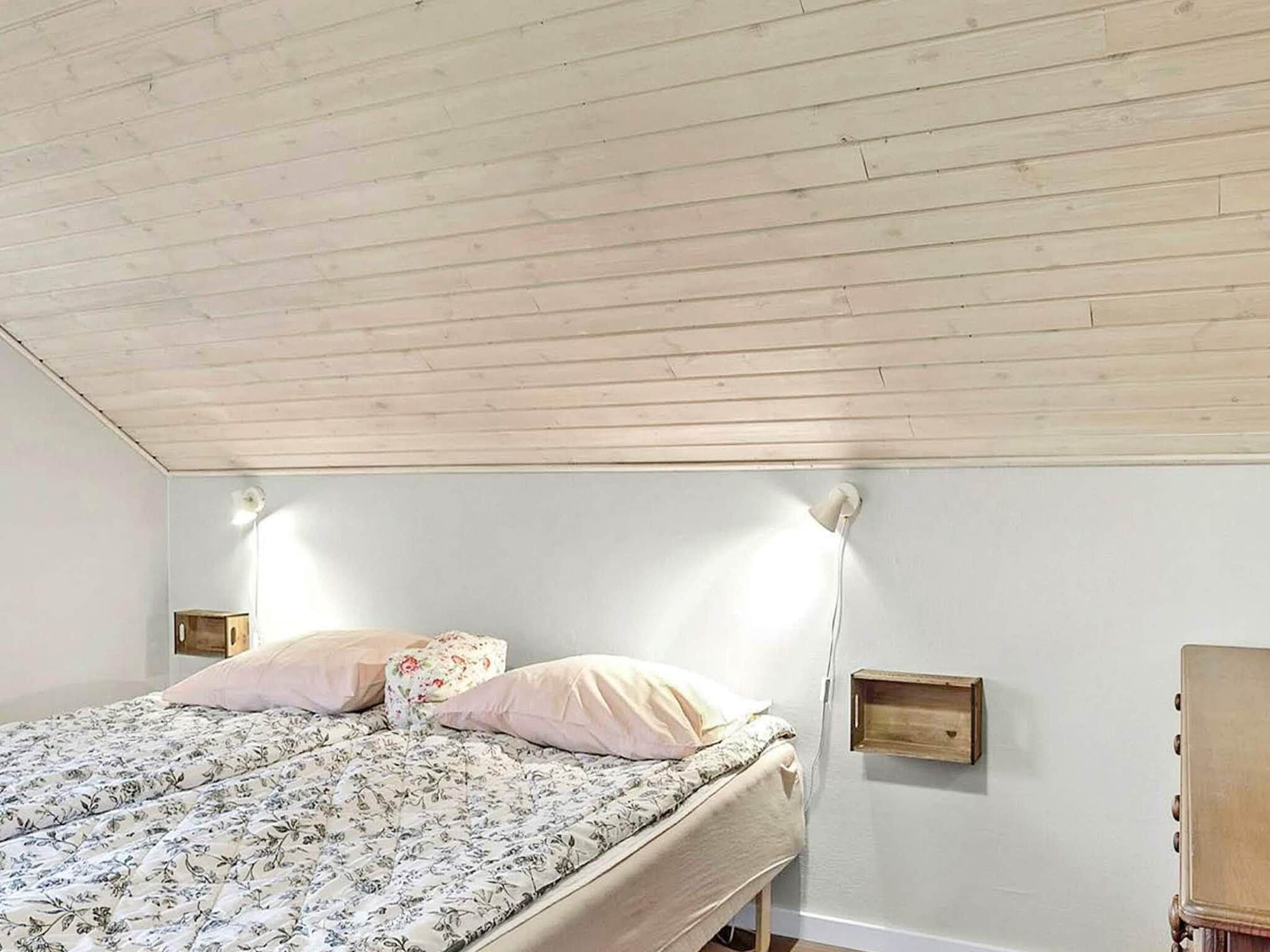 A bedroom with a wooden ceiling, a bed with floral bedding, two bedside tables, and a wall-mounted shelf.