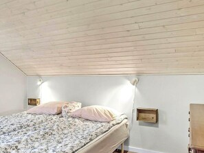 A bedroom with a wooden ceiling, a bed with floral bedding, two bedside tables, and a wall-mounted shelf.