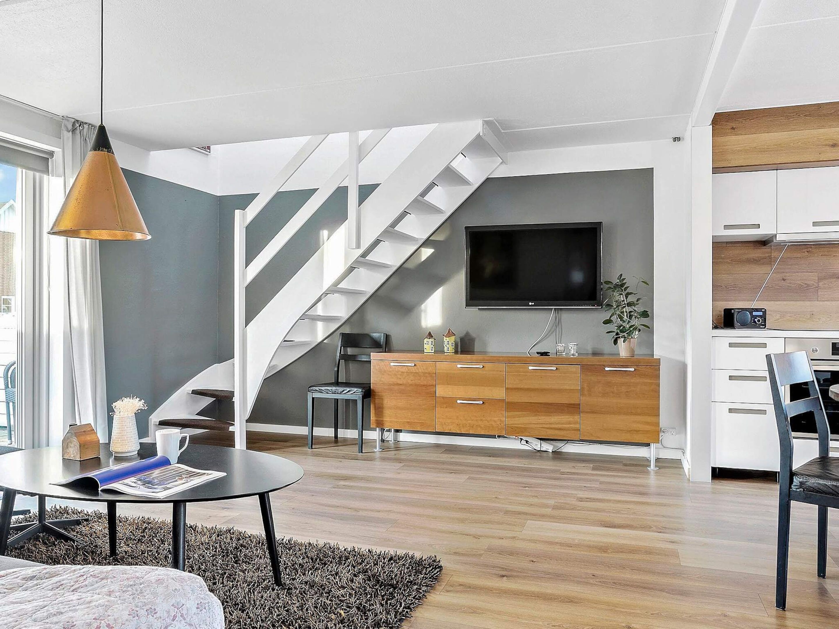 A modern living room with a staircase, a flat-screen TV, wooden cabinets, and a dining area.