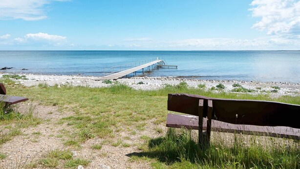 Ein Strand mit einer hölzernen Pier, die sich über ruhiges Wasser erstreckt, einer Rasenfläche mit einer Bank und einem klaren Himmel.