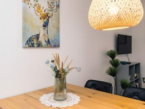 A dining area with a wooden table, a vase with dried plants, a painting of a deer, a woven light fixture, and a television on a stand. - 6 Person Holiday Home in Hadsund (Hadsund)