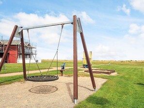 A swing set in a park with a red building in the background. - 6 Person Holiday Home in Hadsund (Hadsund)