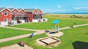 A red building with a balcony, a playground with a blue play structure, and a clear view of the beach. - 6 Person Holiday Home in Hadsund (Hadsund)