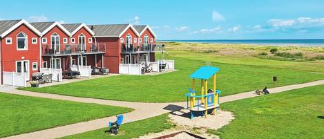 A red building with a balcony, a playground with a blue play structure, and a clear view of the beach.