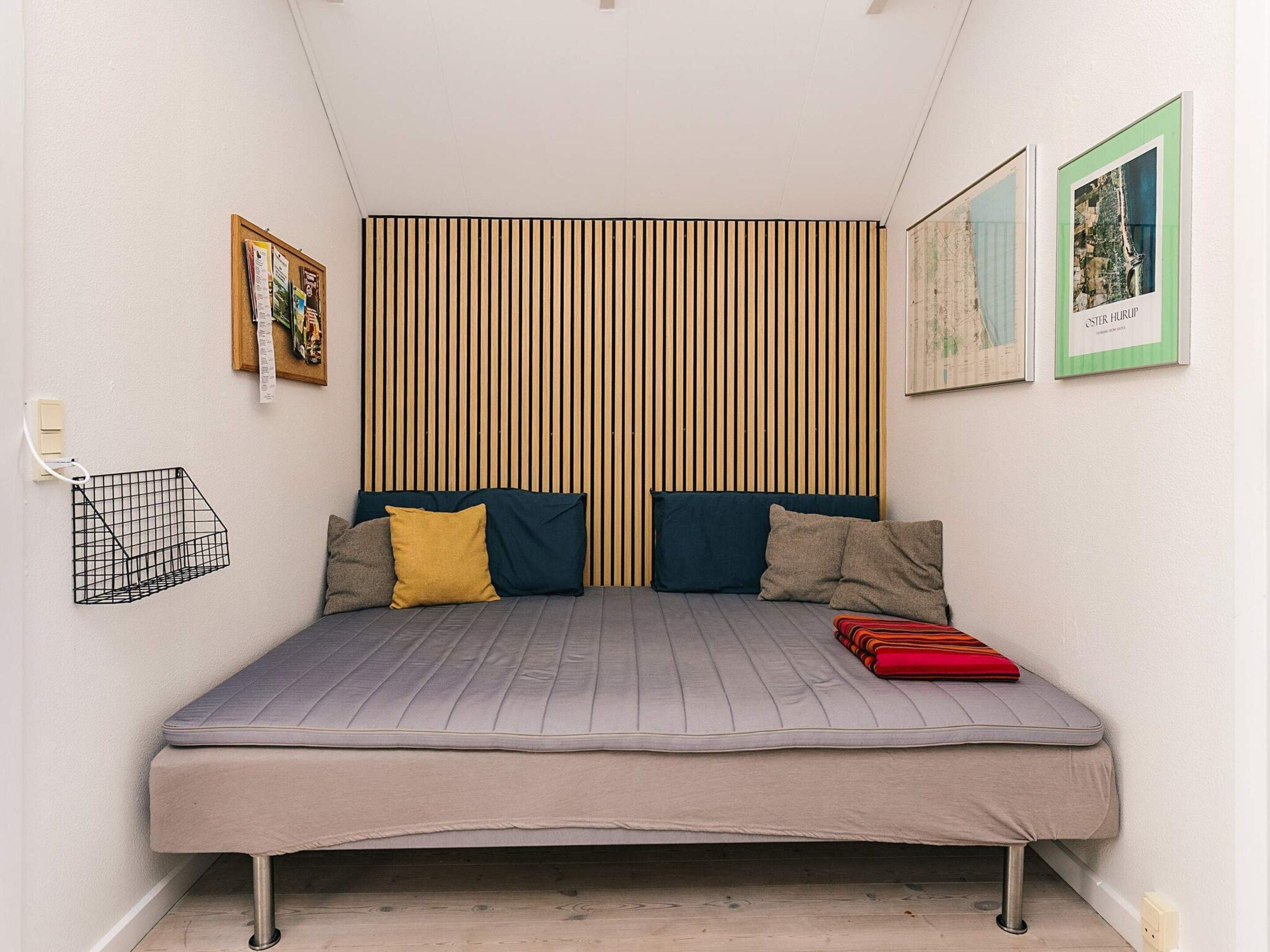 A small, neatly arranged bedroom with a single bed, a slatted headboard, a small shelf, and two framed pictures on the wall.