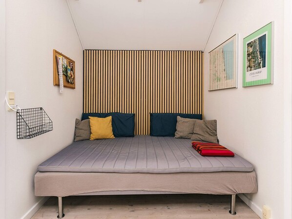 A small, neatly arranged bedroom with a single bed, a slatted headboard, a small shelf, and two framed pictures on the wall.