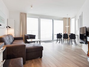 A modern living room with a sofa, a dining area with chairs and a table, a television, and large windows with curtains. - 5 Star Holiday Home in Wendtorf (Wendtorf)