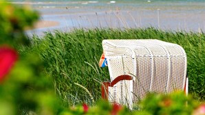 Eine Strandansicht mit einem weißen Liegestuhl im Vordergrund, einem Sandstrand und dem Meer im Hintergrund.
