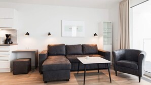 Ein modernes Wohnzimmer mit einer Couch, einem Couchtisch und einem Sessel. Es gibt eine Küchenzeile mit einer Kaffeemaschine und einem Schrank.