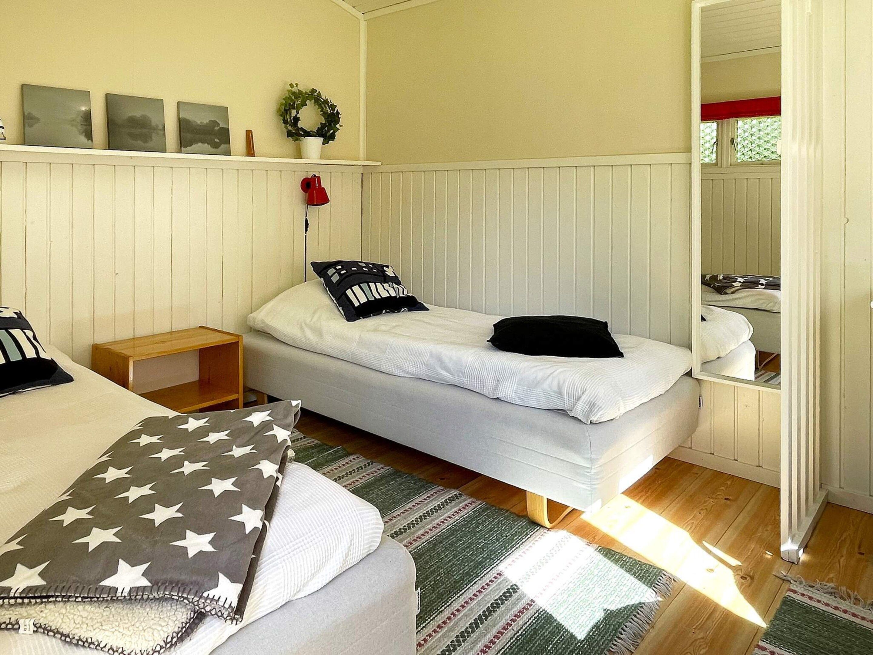 A single bed with a white mattress and pillows, a wooden nightstand, a small table, a wall-mounted shelf with decorative items, and a window with a view of greenery.