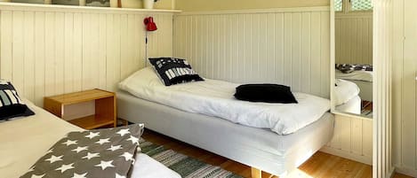 A single bed with a white mattress and pillows, a wooden nightstand, a small table, a wall-mounted shelf with decorative items, and a window with a view of greenery.