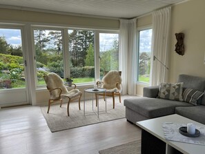 Een kamer met een grijze bank, twee fauteuils, een kleine tafel en uitzicht op een tuin door grote ramen.