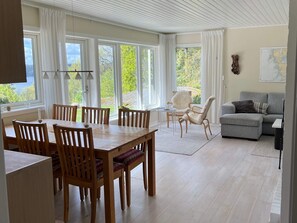 A spacious living area with a dining table, chairs, a sofa, and a view of greenery through large windows.
