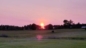 Auringonlasku niityn yllä, taustalla näkyy puita ja kauempana pieni rakennus.