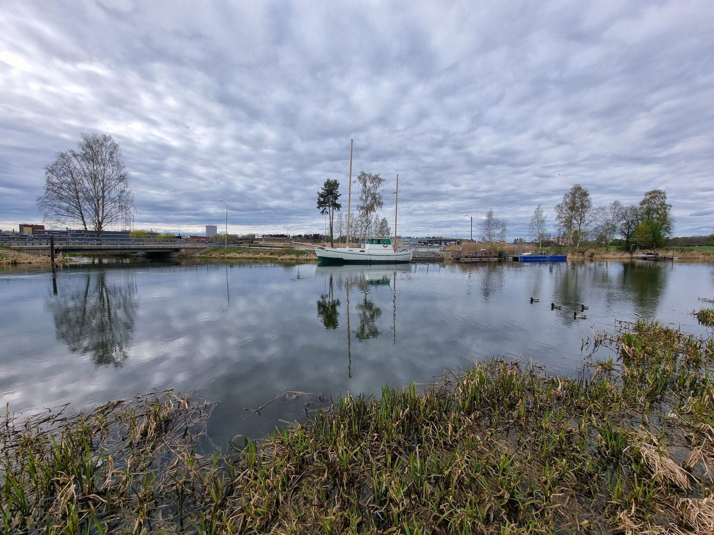 A calm river with boats docked, surrounded by trees and a cloudy sky.