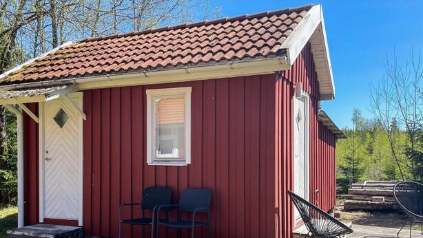 Une cabane en bois rouge avec un toit en tuiles, une porte blanche, deux chaises et une petite fenêtre.