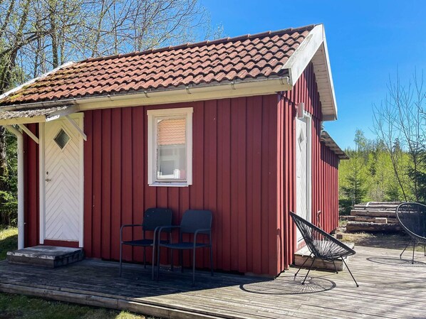 A red wooden cabin with a tiled roof, a white door, two chairs, and a small window.