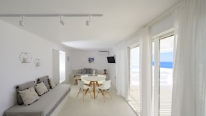 Uma sala de estar moderna com sofá, mesa de jantar e televisão. Há uma janela grande com cortinas e vista para a praia.