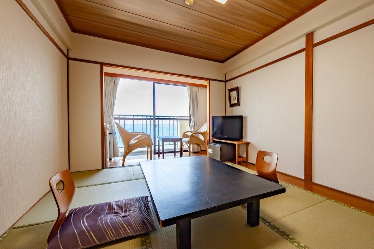 A room with tatami flooring, a low table, chairs, a television, and a balcony with a view.