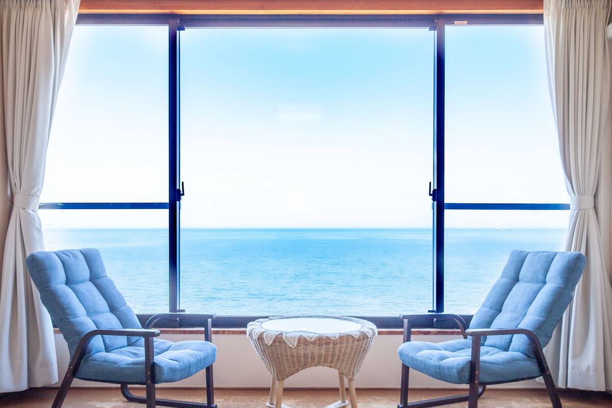 A room with two blue armchairs and a small wicker table, a large window with a view of the sea, and curtains on either side.