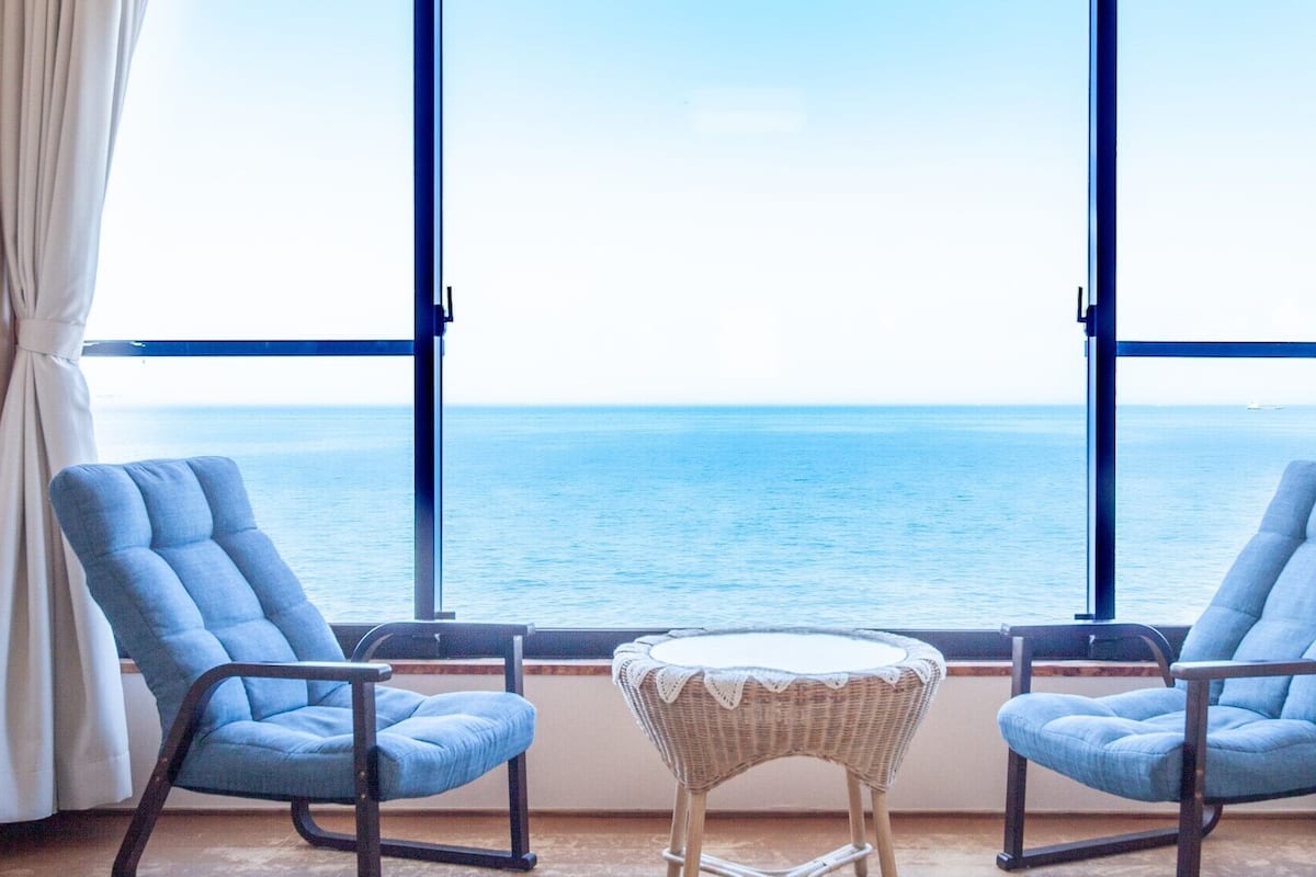 A room with two blue armchairs facing a large window overlooking the sea.
