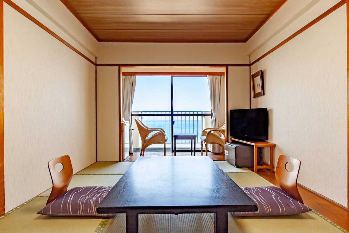 A room with a wooden ceiling, a large table, two chairs, a television, and a view of the sea through a sliding door.