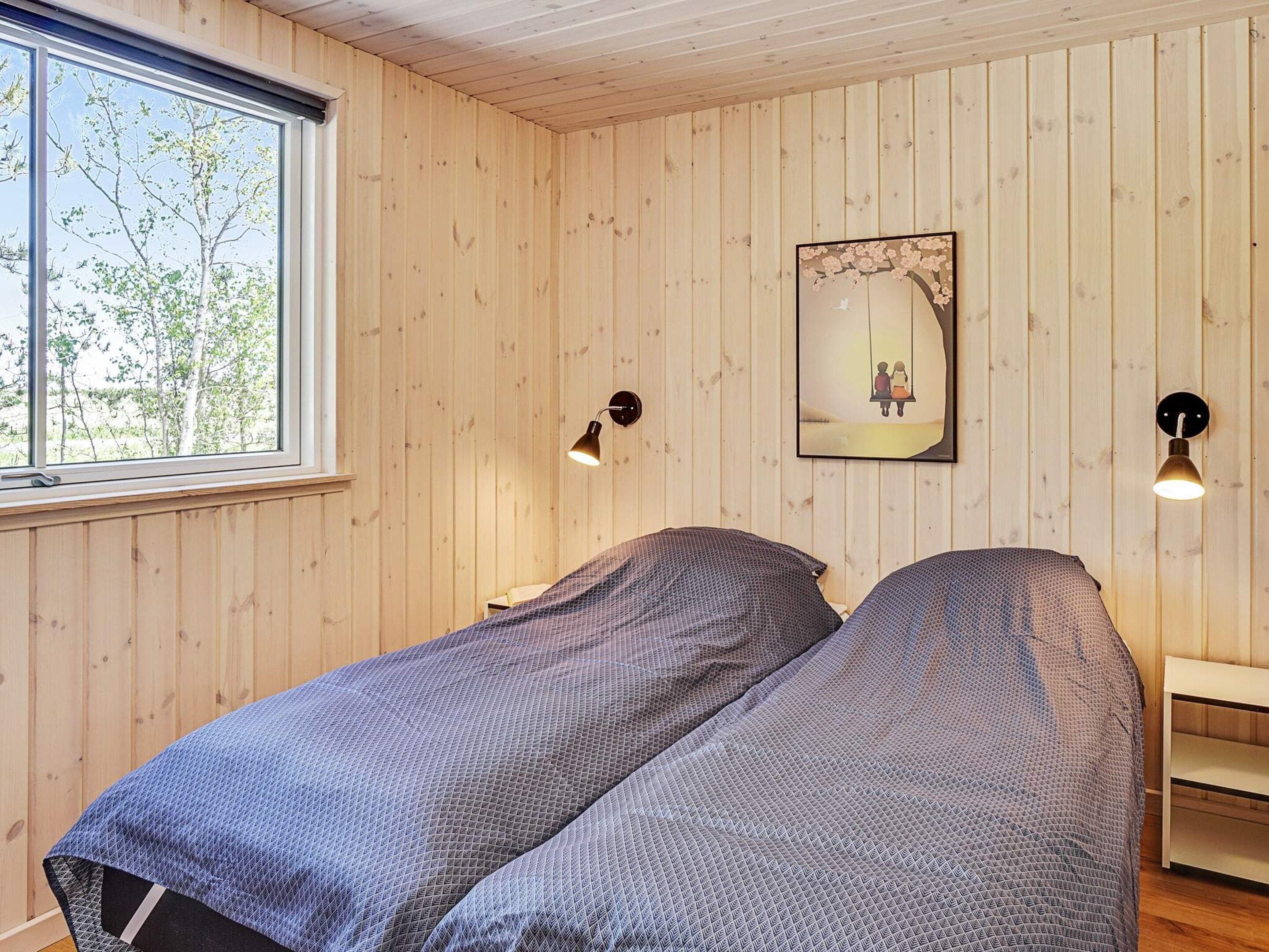 A room with wooden walls, a bed with a patterned bedspread, a window with a view of trees, and wall-mounted lamps.