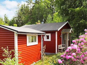 A red cabin with a small porch and a bench, surrounded by lush greenery and pink flowers.