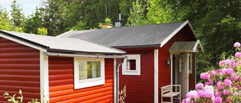 A red cabin with a small porch and a bench, surrounded by lush greenery and pink flowers.