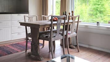Un comedor con mesa y sillas de madera, una ventana con vistas a follaje, y una cocina con gabinetes blancos.