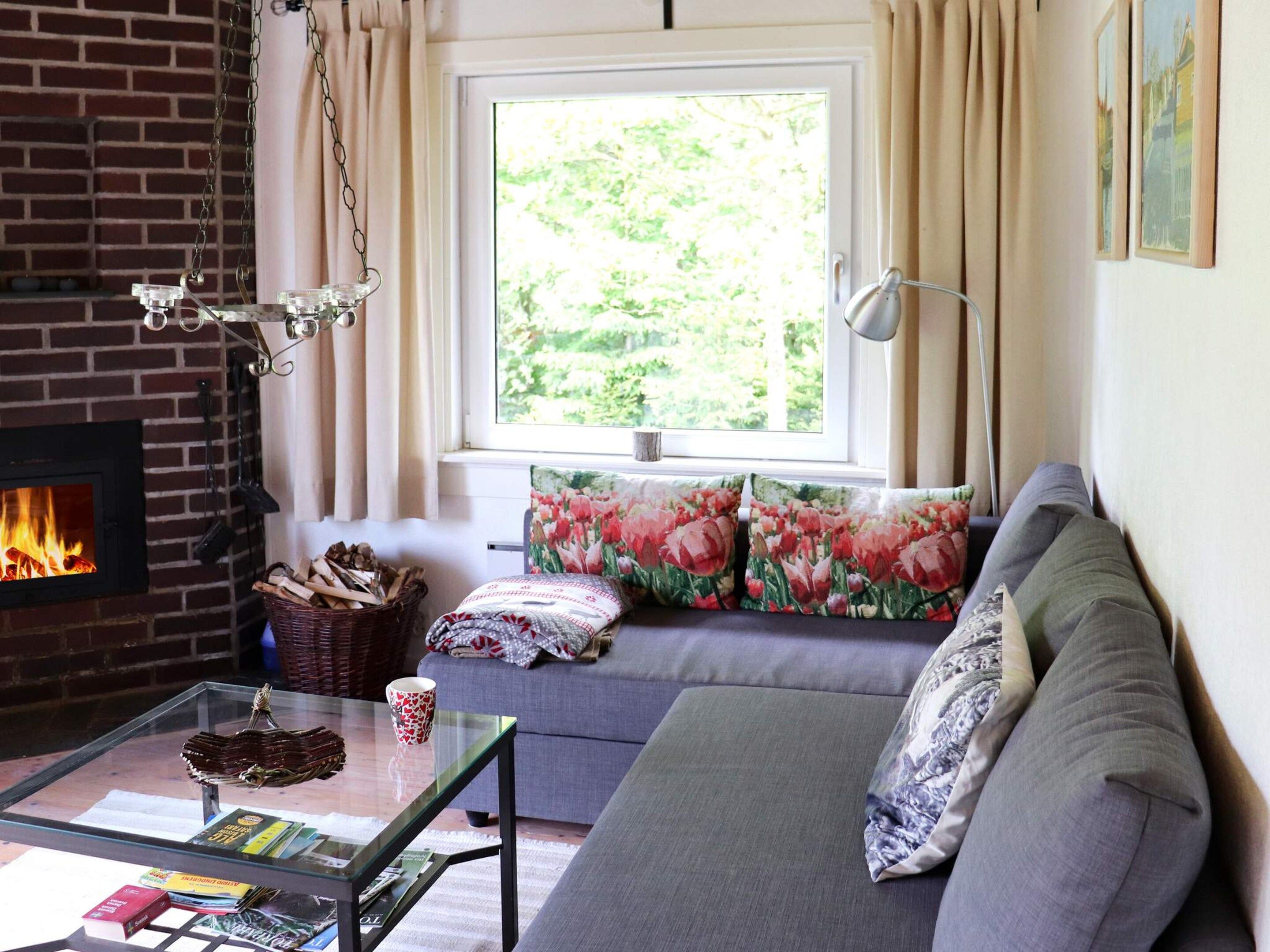 Una sala de estar con chimenea, un sofá con cojines florales, una mesa de centro de vidrio con libros y una taza, y vistas a la vegetación a través de una ventana.