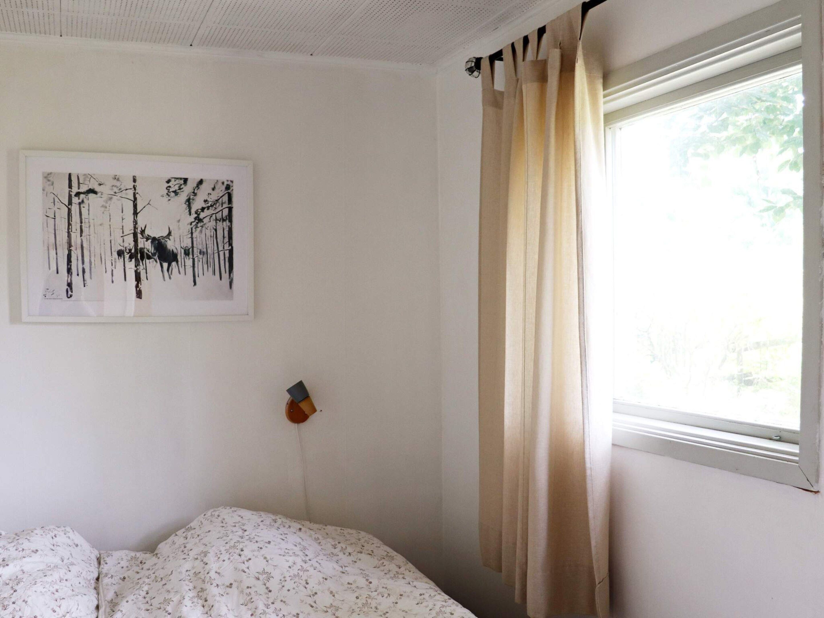 Un dormitorio con cama, lámpara de pared, ventana con cortinas e imagen en el marco colgada en la pared.