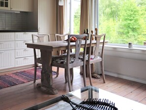 A dining area with a wooden table and chairs, a window with a view of greenery, and a kitchen with white cabinets.
