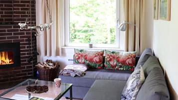 Una sala de estar acogedora con chimenea, un sofá con cojines estampados, una mesa de centro de vidrio con libros y vistas al verdor exterior.