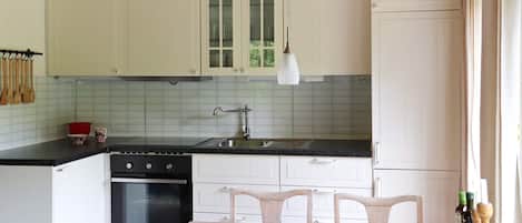 A kitchen with white cabinets, a black countertop, a sink, and a dining table with chairs. There are wooden utensils hanging on the wall and a bowl of fruit on the table.