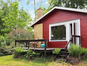 A red cabin with a brick chimney, a deck with a table and chairs, and a window overlooking the surrounding greenery.