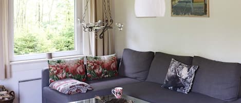 Een woonkamer met een grijze bank, een glazen salontafel, een hanglamp, een raam met gordijnen en een schilderij aan de muur.