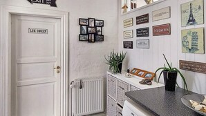 A kitchen with a white door labeled "LUK DØREN," a radiator, a countertop with a plant, and various framed signs on the wall.
